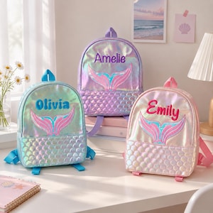 Op de afbeelding: Drie iriserende rugzakken met zeemeerminstaartborduursel en gepersonaliseerde namen. De linker tas is blauw met de naam Olivia, het midden is paars met Amelie en de rechter is roze met Emily. Elke tas heeft een voorvak met schubbenpatroon.