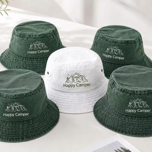 Pode incluir: Quatro chapéus de pescador verde-escuros e um chapéu branco, cada um com um gráfico bordado de linhas de montanhas, um sol e o texto Happy Camper. Os chapéus têm um topo plano, uma aba inclinada e ilhós metálicos nas laterais.