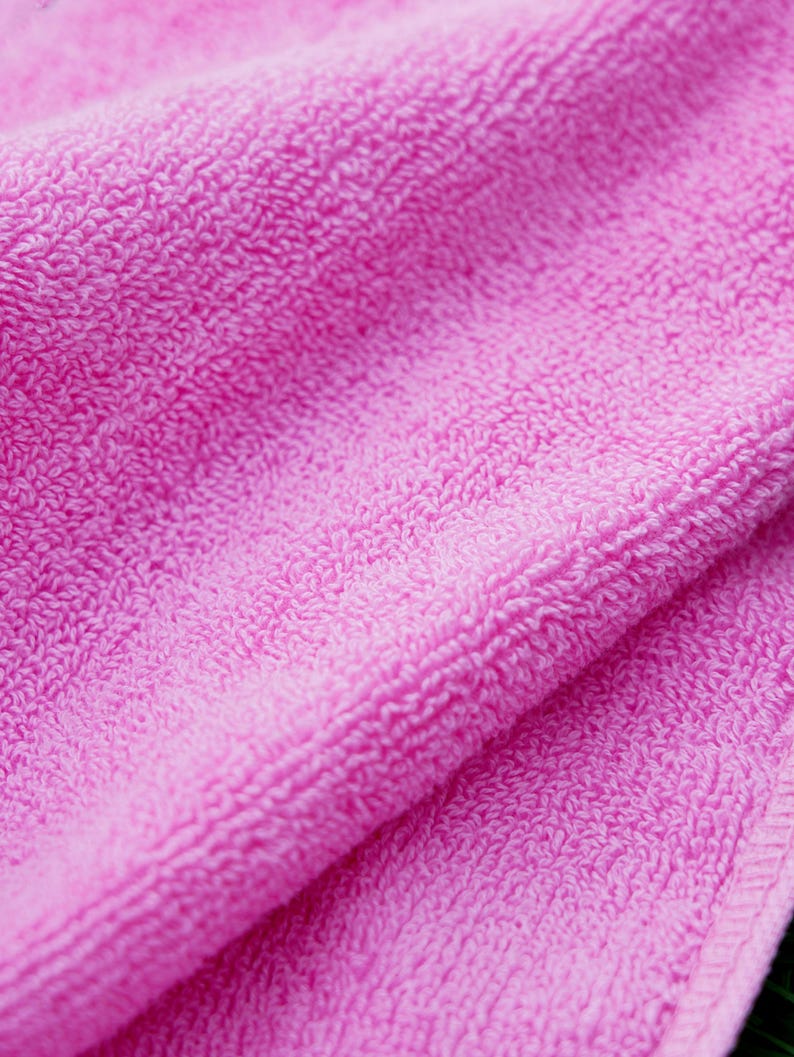 May include: Close-up of a folded, bright pink terry cloth towel. The soft, textured surface of the towel is visible, with a detailed view of the looped fibres. The edges of the towel are neatly finished.
