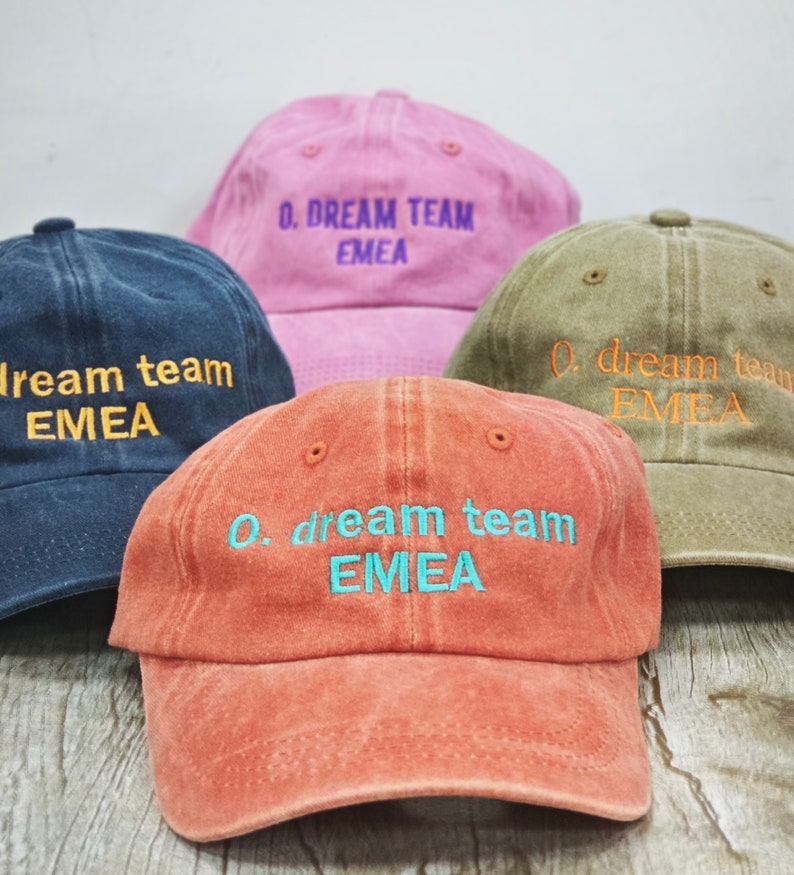 K&ouml;nnte beinhalten: Vier Baseballkappen in verschiedenen Farben mit dem gestickten Text "0. dream team EMEA" auf der Vorderseite. Die Kappen sind aus weichem, strukturiertem Stoff.