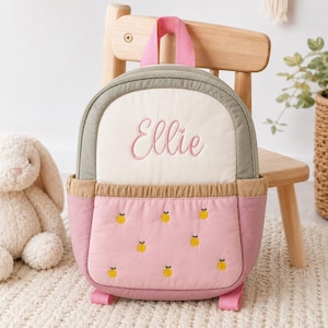 Può includere: Uno zaino multicolore con una sezione superiore color crema con il nome Ellie ricamato in rosa, un bordo verde salvia e una sezione inferiore rosa decorata con piccoli motivi di arance gialle ricamate e una tasca elastica color sabbia.