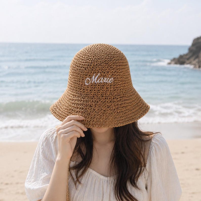 May include: A woven straw sun hat with a wide brim, embroidered with the name "Marie" in white script. The hat is a light brown color and is being held up by a person on a beach with the ocean in the background.