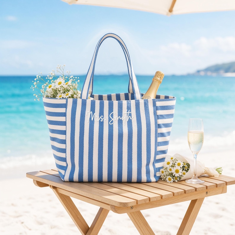 May include: A blue and white striped tote bag with two handles, featuring the white embroidered text "Mrs. Smith" on the front. The bag is filled with white daisies and a champagne bottle, sitting on a wooden table near the ocean.