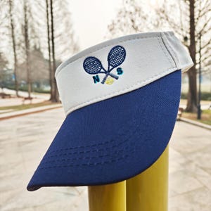May include: A navy blue and white tennis visor. The visor has an embroidered design of crossed tennis rackets, a tennis ball, and the letters "N" and "S". The visor is made of a durable fabric and is designed to shield the eyes from the sun.
