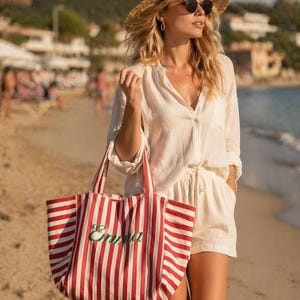 May include: A large tote bag featuring vertical red and white stripes with the name Emma embroidered in green cursive script on the front. The bag has two matching striped fabric handles and a spacious open-top design for carrying items.