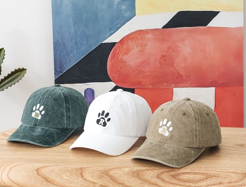 K&ouml;nnte beinhalten: Drei Baseballkappen mit Pfotenabdruck-Designs. Die Kappen sind gr&uuml;n, wei&szlig; und braun. Die Pfotenabdr&uuml;cke haben einen Buchstaben in der Mitte.