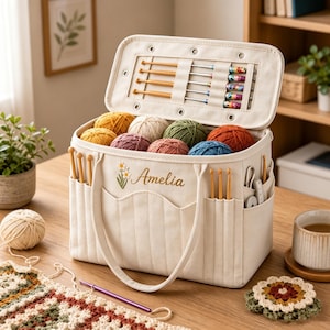 May include: A cream-colored canvas yarn storage bag with the name Amelia embroidered in brown script next to a small daisy graphic. The bag features multiple exterior pockets holding crochet hooks, an open top filled with colorful yarn balls, and a lid with internal elastic loops for additional crochet hooks.