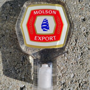 May include: A clear, hexagonal tap handle with the Molson Export logo. The logo features a red and white design with a blue ship and the words "Molson" and "Export". The handle has a clear, cylindrical base.