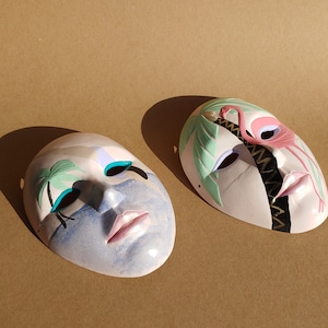 Peut inclure: Deux masques en céramique avec des motifs peints à la main. Un masque représente une scène tropicale bleue et blanche avec des palmiers. L'autre masque représente un flamant rose avec des palmiers verts et un motif en zigzag noir et or.