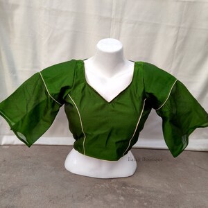 May include: A green blouse with a V-neck and short, flared sleeves. The blouse has a gold trim around the neckline and sleeves. The text "Balaji Boutique" is visible in the bottom right corner of the image.