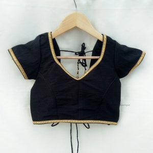 May include: Black cotton blouse with gold trim and a tie back. The blouse has short sleeves and a V-neck.