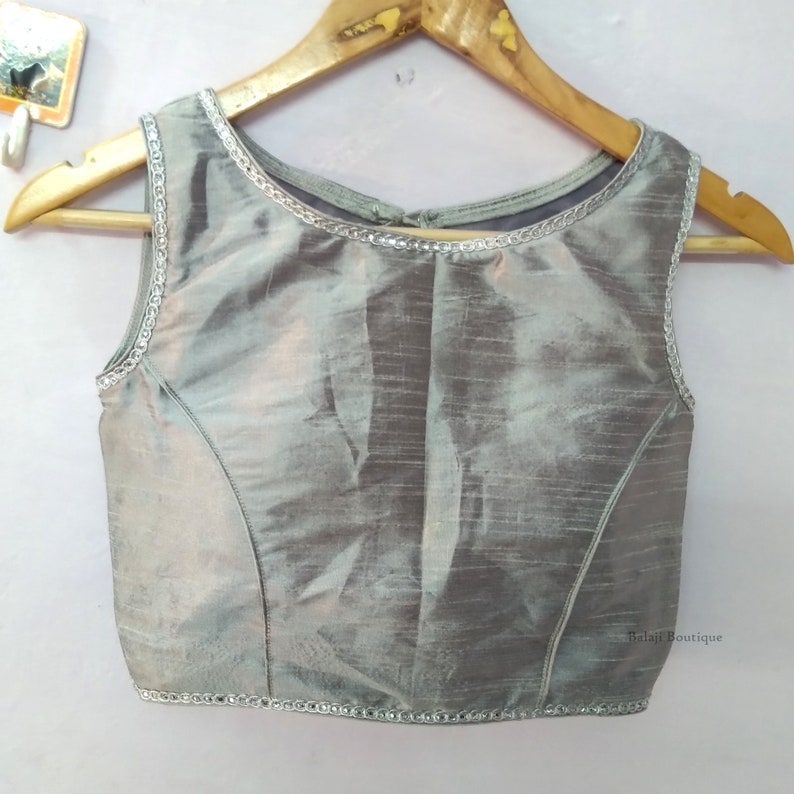 May include: Sleeveless silver-gray blouse with a round neckline and a subtle sheen. The edges are trimmed with a delicate silver embellishment. The blouse is hanging on a wooden hanger.
