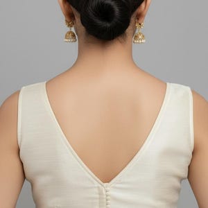 May include: A sleeveless off-white blouse featuring a deep V-neckline on the back, accented with a vertical row of small matching fabric buttons running down the center. The garment is paired with gold-toned bell-shaped drop earrings.