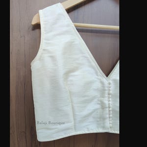 May include: A sleeveless off-white blouse with a deep V-neckline and a vertical row of small fabric-covered buttons along the front closure. The garment features a subtle textured finish and includes the text Balaji Boutique on the lower left.