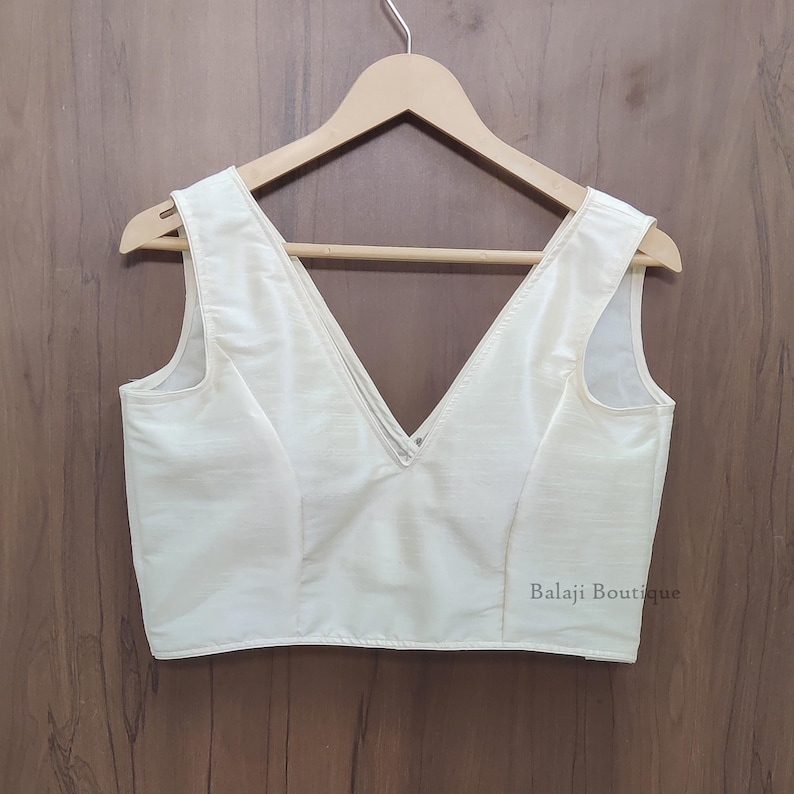 May include: A sleeveless off-white blouse featuring a deep V-neckline and a structured bodice. The garment is displayed on a wooden hanger and includes the text Balaji Boutique printed in small black letters on the lower right side of the fabric.