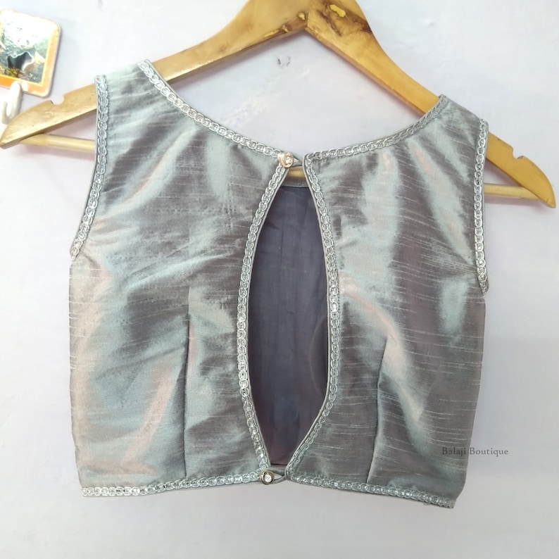 May include: Sleeveless silver blouse with a keyhole back. The blouse has a subtle sheen and is trimmed with a delicate silver border. A small, decorative button secures the back opening. The blouse is displayed on a wooden hanger.