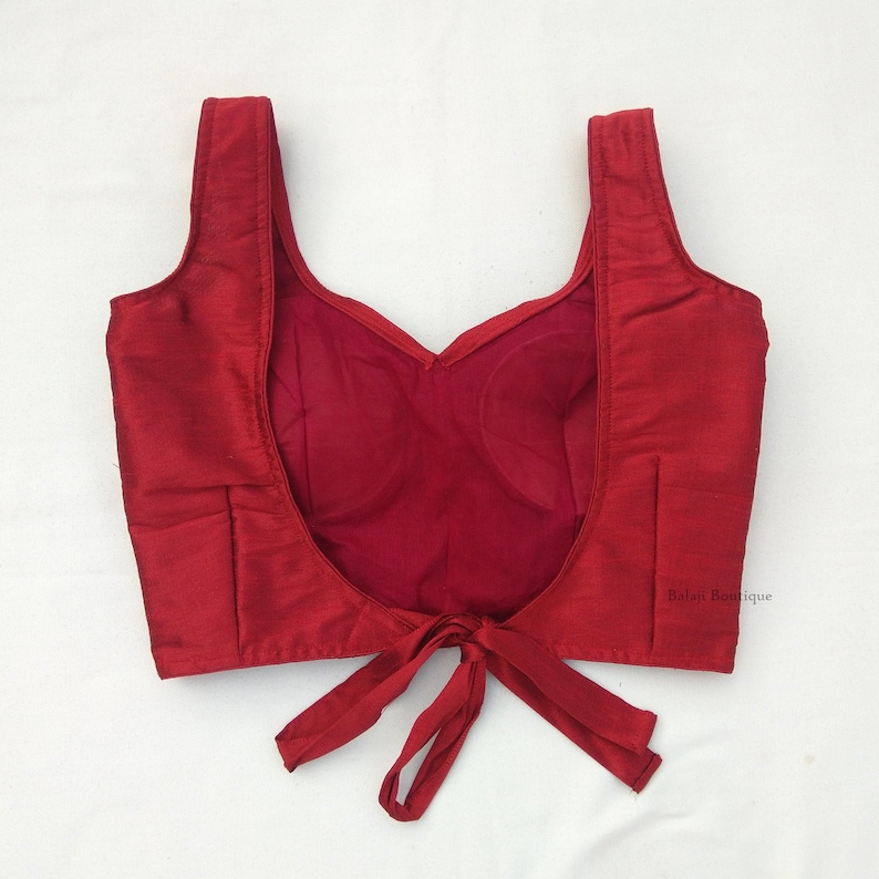 May include: A maroon sleeveless blouse with a deep V-shaped back and a tie closure at the bottom. The blouse is made of a smooth, silky fabric.