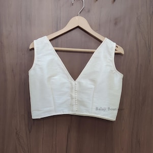 May include: A sleeveless off-white blouse featuring a deep V-neckline and a vertical row of small matching fabric-covered buttons down the center front. The garment is displayed on a wooden hanger and includes the text Balaji Boutique.