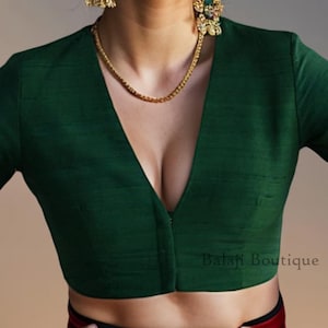 May include: A dark green blouse with long sleeves and a deep V-neck. The blouse has a simple, elegant design and is made of a smooth fabric. A gold necklace and earrings are visible. The text "Balaji Boutique" is printed on the bottom right.