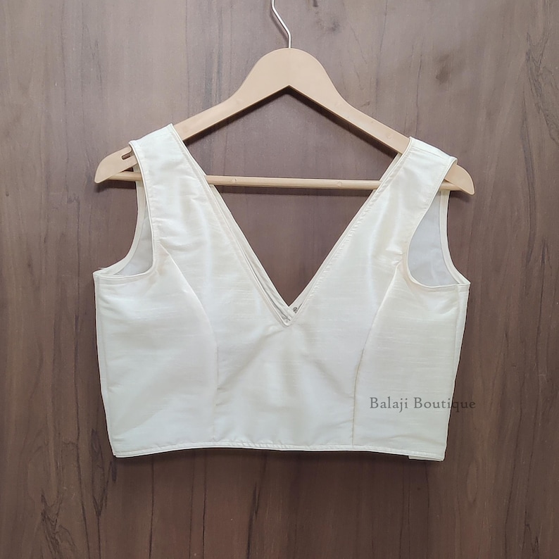 May include: A sleeveless, off-white blouse featuring a deep V-neckline and a structured bodice. The garment is displayed on a wooden hanger and includes the text Balaji Boutique printed in small, dark letters on the lower right side.