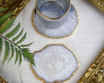 May include: Two agate coasters with gold edging and a clear glass with a gold rim. The coasters are white and light blue with a natural, organic pattern. The coasters are on a white surface.