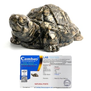 May include: A metallic pyrite tortoise sculpture. The sculpture is a dark color with gold flecks and is sitting on a white surface. A lab report card is in front of the sculpture, with the text "NATURAL PYRITE".