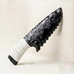 May include: A black obsidian knife with a serrated edge. The handle is wrapped with white cord. The blade has a textured surface. The knife is displayed against a neutral background.