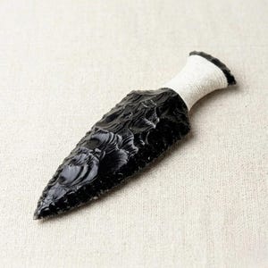 May include: A handcrafted obsidian arrowhead with a textured, glossy black surface. The handle is wrapped with white cord. The arrowhead is approximately 15 cm long.