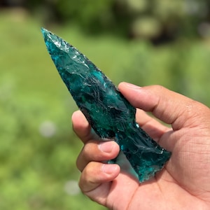 May include: A hand holding a teal-colored glass arrowhead. The arrowhead has a sharp point and a textured surface. The background is blurred green foliage.