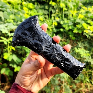 May include: A black obsidian arrowhead with a sharp point and a smooth, polished surface. The arrowhead is held in a hand against a green background.