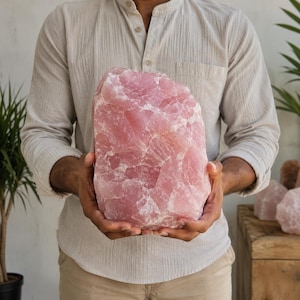 Può includere: Un grande pezzo grezzo di quarzo rosa con una tonalità rosa pallido traslucida e una consistenza cristallina irregolare. La pietra presenta fratture naturali e variazioni di opacità, mostrando una superficie morbida che riflette la luce.