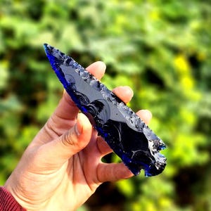 May include: A hand holds a dark blue obsidian arrowhead against a blurred green background. The arrowhead is a deep, translucent blue at the edges, transitioning to black. The edges are sharp and jagged, with a classic arrowhead shape.
