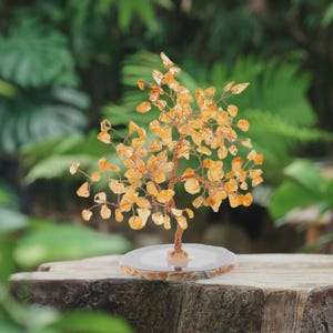 Può includere: Una scultura decorativa a forma di albero con un tronco e rami in filo di rame, adornata con foglie di pietre preziose giallo-arancio. L'albero poggia su una base rotonda di colore chiaro, su uno sfondo verde sfocato. La scultura è alta circa 15 cm.