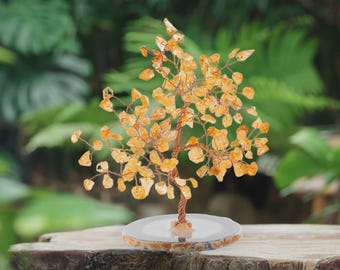 May include: A decorative tree sculpture with a copper wire trunk and branches, adorned with orange-yellow gemstone leaves. The tree sits on a round, light-colored base, set against a blurred green background. The sculpture is approximately 6 inches tall.