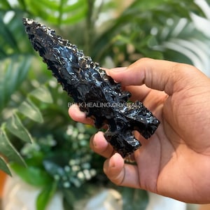 May include: A black obsidian arrowhead with a textured surface. The arrowhead is held in a hand, with the text "BULK HEALING CRYSTALS" visible. The background features green foliage.