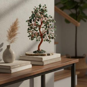 May include: A decorative bonsai tree with a copper-colored trunk and green gemstone leaves sits atop a stack of books on a wooden table. A small vase with dried pampas grass and a potted plant are also on the table.
