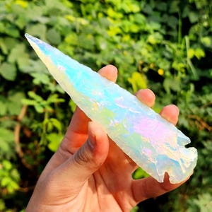 May include: A large, iridescent, white crystal point with a pointed tip. The crystal has a rainbow sheen and is held in a hand.