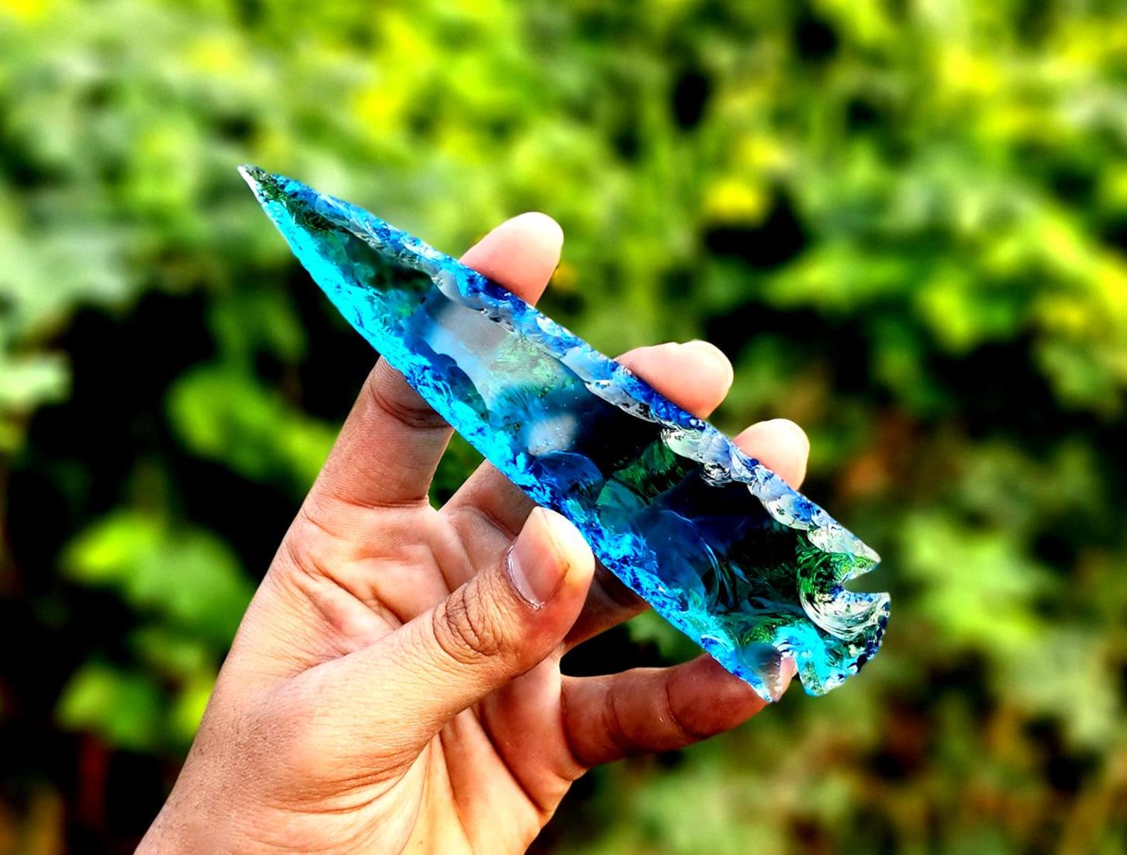 Blue obsidian arrowhead with a reflective surface, held in hand against a blurred green background to enhance Aquarius' mental clarity.