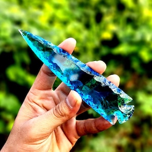 May include: A blue and green glass arrowhead with a sharp point and a jagged edge.