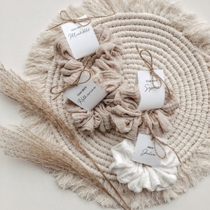Könnte beinhalten: Vier weiße Stoffscrunchies mit befestigten Etiketten. Die Etiketten sind weiß mit schwarzem Text. Der Text auf den Etiketten lautet "Team Bride", "Bride", "Team Bride" und "Team Bride". Die Scrunchies liegen auf einer beigefarbenen gewebten Matte mit Fransenrand.