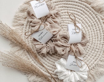 May include: Four white fabric scrunchies with tags attached. The tags are white with black text. The text on the tags reads "Team Bride", "Bride", "Team Bride", and "Team Bride". The scrunchies are on a beige woven mat with a fringe border.