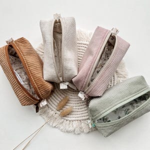 Könnte beinhalten: Vier verschiedene farbige Reißverschlusstaschen mit einem floralen Futter. Die Taschen sind aus weichem Cordstoff gefertigt. Die Taschen sind auf einer weißen, gehäkelten Oberfläche angeordnet.