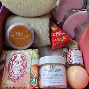 May include: A pink gift box filled with bath and body products. The box contains a white washcloth, a pink eye mask, a bar of soap with a floral design, a jar of Himalayan salt bath soak, a small bottle of strawberry passion car diffuser, and a round, peach-colored bath bomb. The products are all from the Millwood Naturals & Organics brand.
