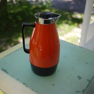 Può includere: Un thermos vintage arancione e nero con manico nero e bordo argentato. Il thermos è cilindrico con la parte superiore arrotondata e un coperchio nero. È appoggiato su una superficie verde chiaro.