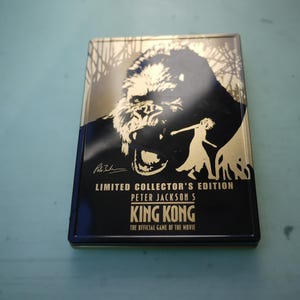 Puede incluir: Edición limitada de coleccionista del juego King Kong de Peter Jackson. La portada presenta un diseño negro y dorado con una silueta de King Kong, una mujer y el texto "King Kong: The Official Game of the Movie."