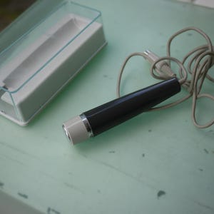 May include: A vintage electric shaver with a black and silver body, a white end, and a coiled gray power cord. The shaver is next to a white and clear plastic case.