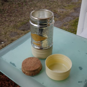 Könnte beinhalten: Ein silberfarbener, strukturierter Metallbehälter mit einem goldfarbenen Emblem, einem beigefarbenen Sockel und einem silbernen Rand. Ein Korken und ein beigefarbener Kunststoffbehälter liegen daneben. Die Artikel befinden sich auf einer hellblauen Oberfläche.