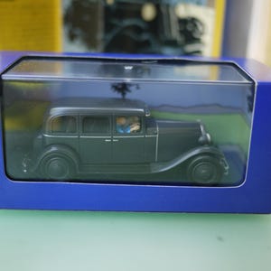 Könnte beinhalten: Ein dunkelgrünes Oldtimer-Modellauto in einer transparenten Kunststoffhülle, in einer blauen Pappschachtel. Das Auto hat ein detailliertes Design, einschließlich Fenster und Räder. Die Box steht auf einer grünen Oberfläche.