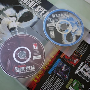 Puede incluir: Se muestran dos CD del videojuego "Rainbow Six: Rogue Spear", junto con el embalaje del juego. Los CD est&aacute;n en fundas de pl&aacute;stico transparente. Un CD es de color rojo oscuro con texto blanco, y el otro es azul y blanco. El embalaje es en blanco y negro.