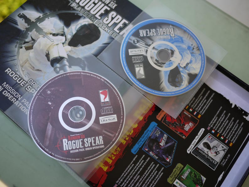 Puede incluir: Se muestran dos CD del videojuego "Rainbow Six: Rogue Spear", junto con el embalaje del juego. Los CD est&aacute;n en fundas de pl&aacute;stico transparente. Un CD es de color rojo oscuro con texto blanco, y el otro es azul y blanco. El embalaje es en blanco y negro.